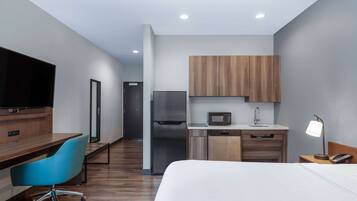 A modern hotel room with a bed, a desk with a chair, a kitchenette with a microwave, refrigerator, and sink, and a television.