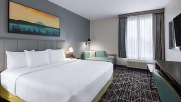 Habitación de hotel con una cama grande, un escritorio, una silla, un sofá, un cuadro en la pared y un ventanal con cortinas.