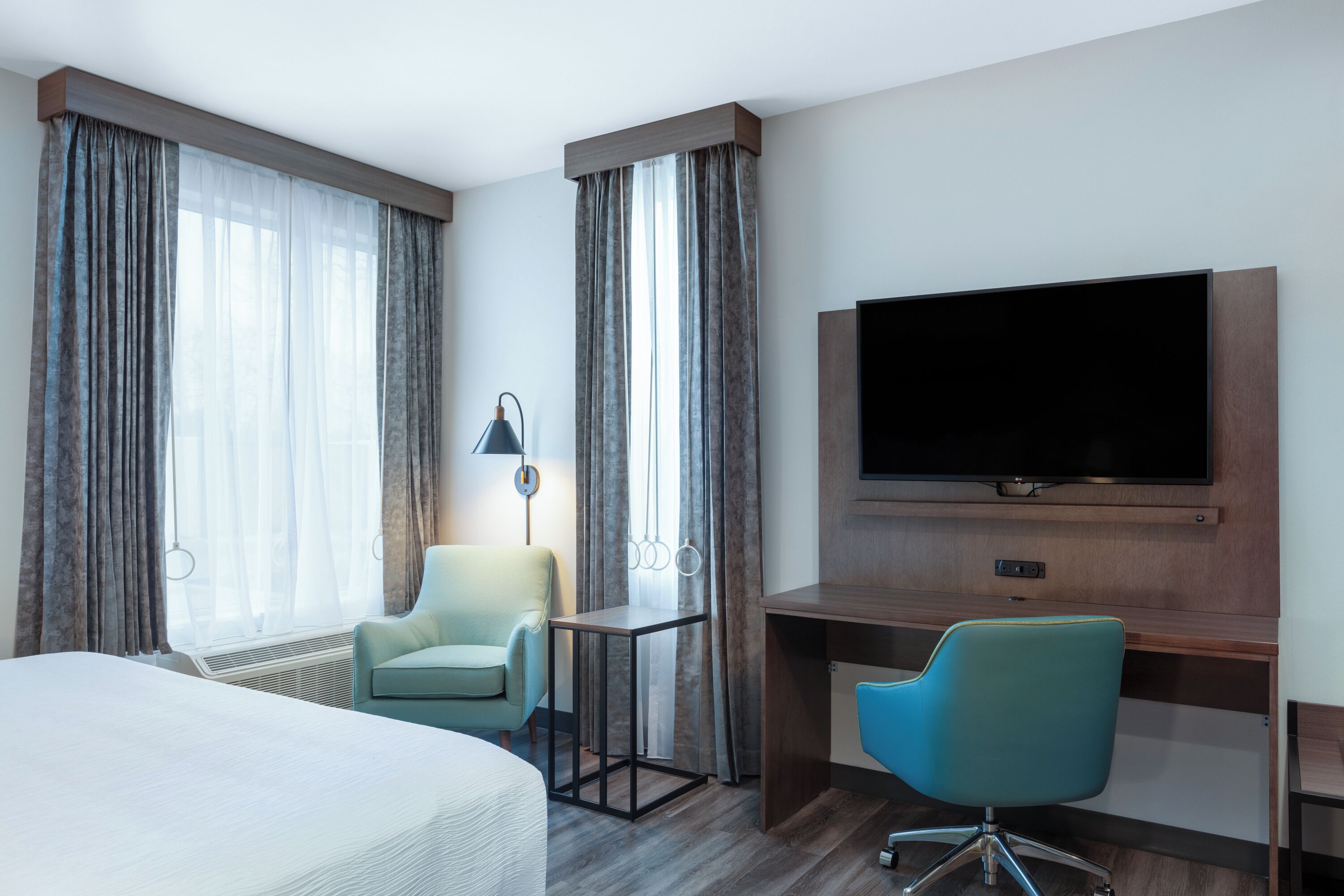 A hotel room with a bed, two chairs, a desk, a television, and large windows with curtains.