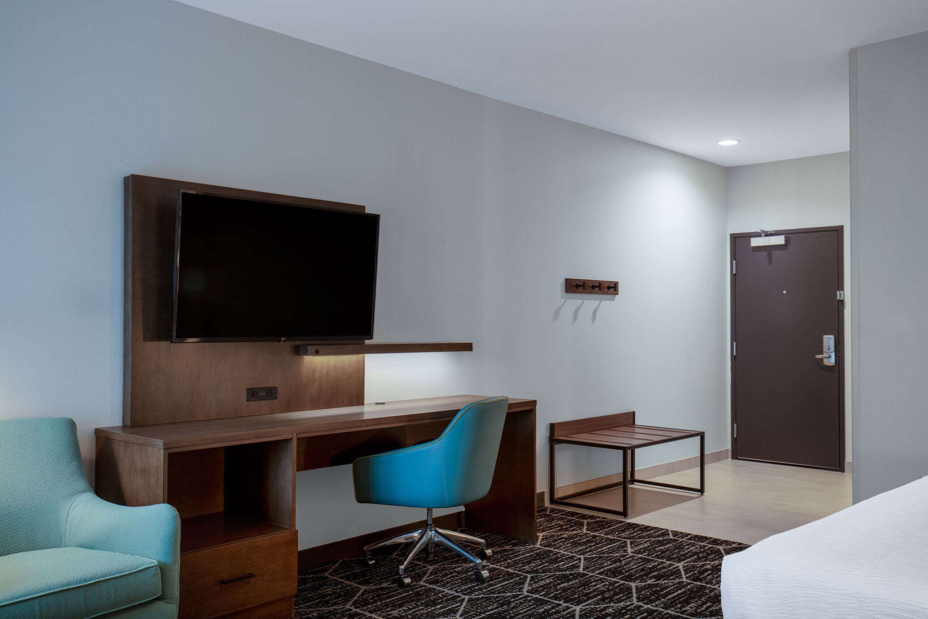 A hotel room with a wooden desk, a blue office chair, a television mounted on the wall, a small table, and a door.