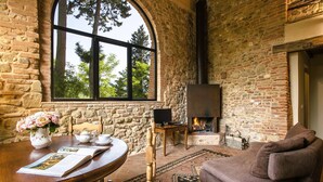 A rustic room with a stone wall, a fireplace, a large window, a round wooden table with a book and teacups, and a brown sofa.