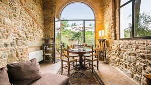 A cozy room with stone walls, a round table with chairs, a sofa, and a large window with an arched view.