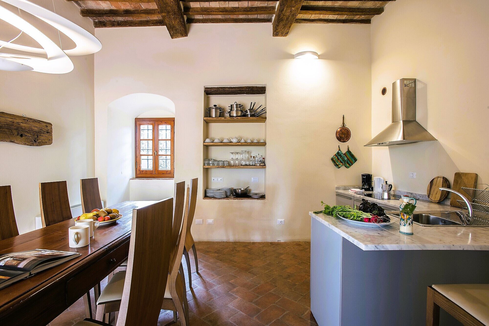 A kitchen with a central island, wooden dining table, and chairs. There is a window with wooden shutters and a shelf with kitchenware.