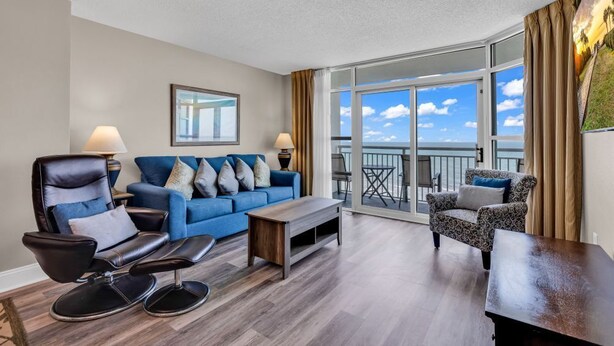 A modern living room with a blue sofa, leather armchair, and patterned armchair. A wooden coffee table is in the center. Large windows offer a view of the sky and clouds. - Breathtaking 2BR Condo w Floor-to-Ceiling Windows Overlooking Ocean (Myrtle Beach)