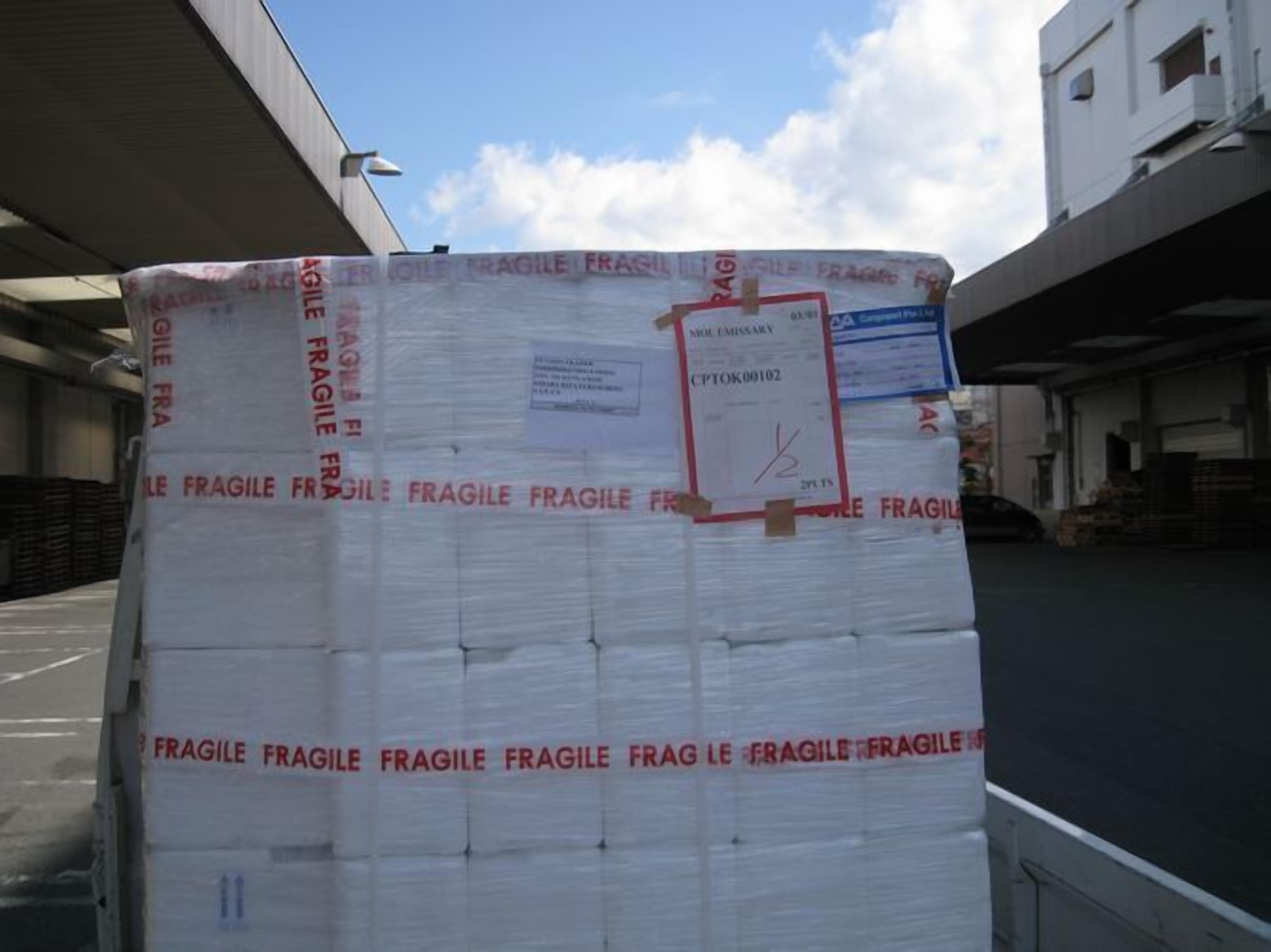 A large, white, rectangular package labeled 'FRAGILE' secured with straps and tape, placed on a flatbed truck in an industrial area with buildings and a clear sky in the background.