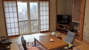 A traditional Japanese-style room with tatami flooring, a low table, a television, and a sliding door with a view of trees.