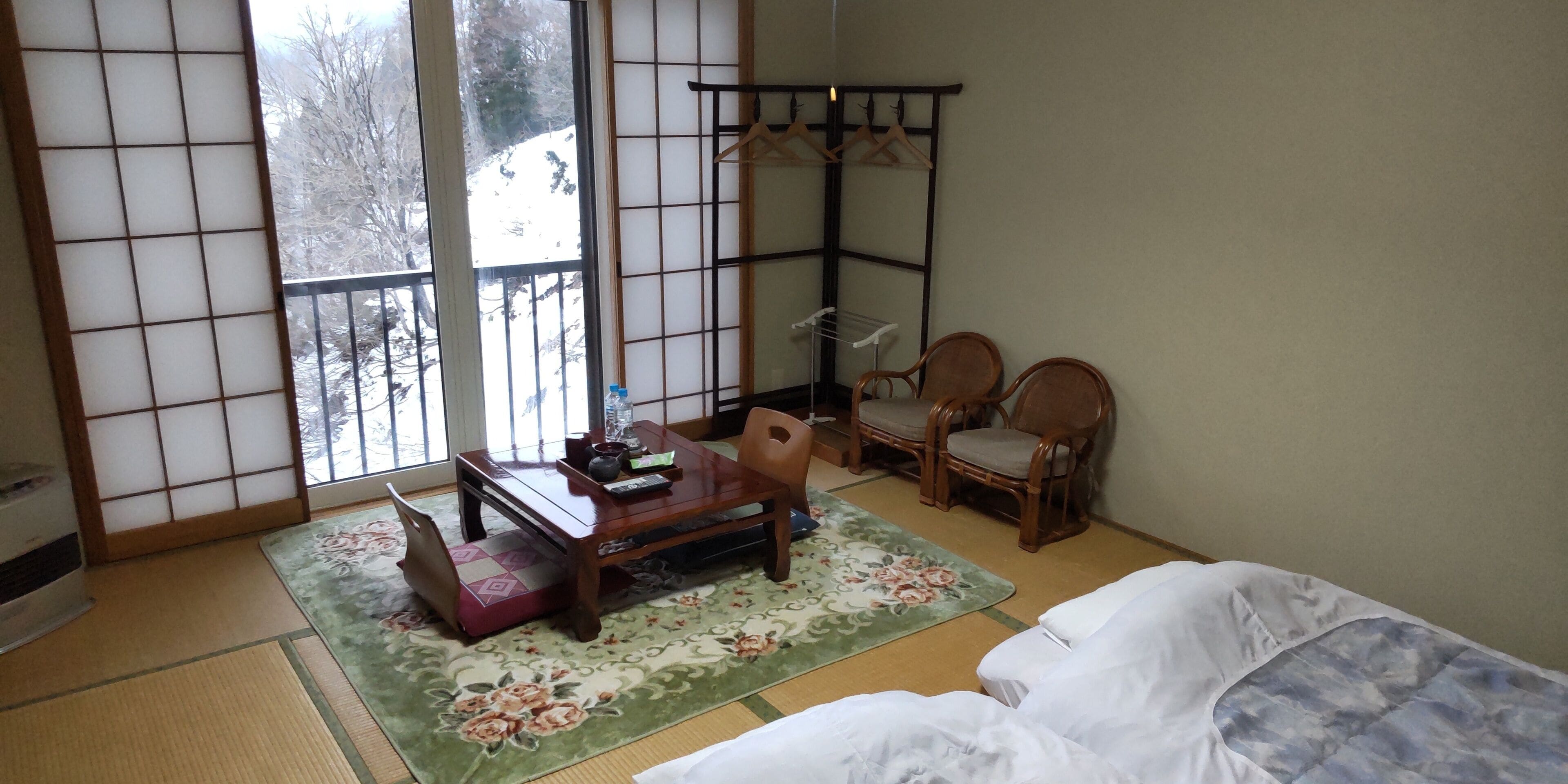 A traditional Japanese room with tatami flooring, a low wooden table, two wooden chairs, a futon bed, a shoji screen, and a view of a snowy landscape.