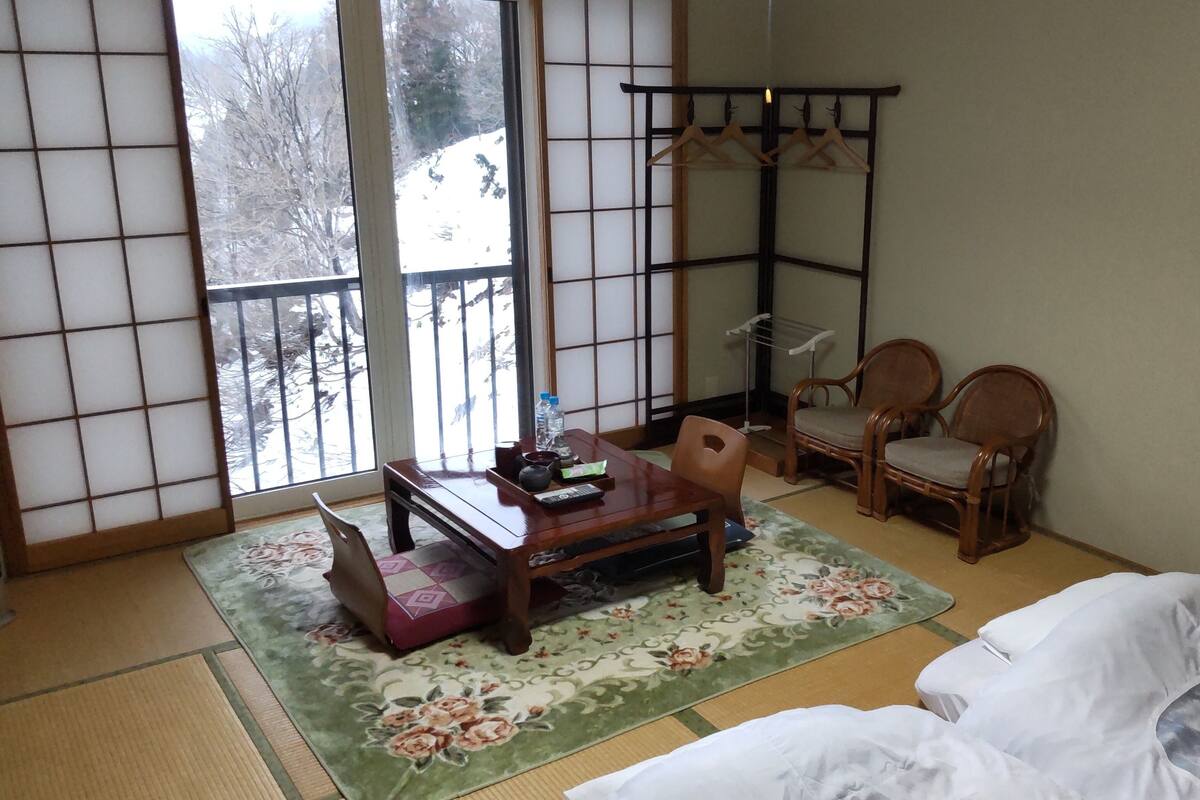 Una habitación japonesa tradicional con suelo de tatami, una mesa de madera baja, dos sillas de madera, un futón y un biombó shoji, con vistas a un paisaje nevado.