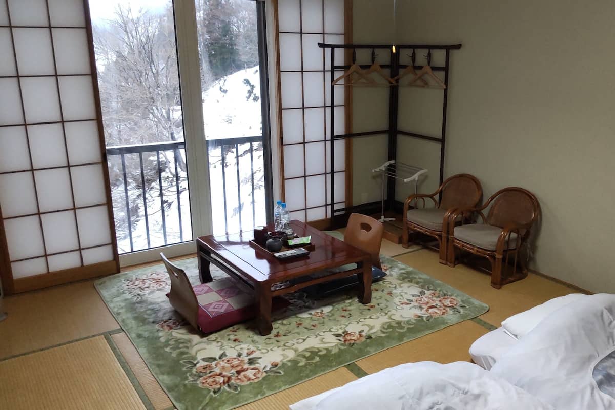 Una habitación japonesa tradicional con suelo de tatami, una mesa de madera baja, dos sillas de madera, un futón y un biombó shoji, con vistas a un paisaje nevado.