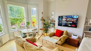 A living room with a beige sofa, a coffee table, and a TV displaying 'THRILLERS'. There are potted plants and a window with a view of greenery.