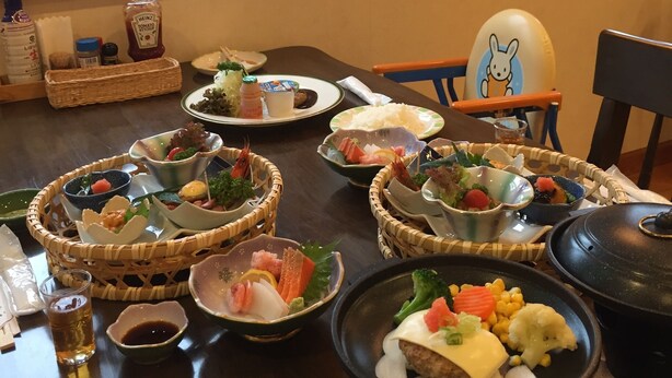 A dining table with various dishes and condiments, a window with a view of greenery, and a framed picture on the wall. - Pension Candle (Fujikawaguchiko)