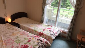 A hotel room with two beds, a bedside table with a lamp, a chair, and a window with curtains. - Pension Candle (Fujikawaguchiko)