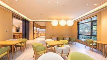 A modern dining area with wooden tables and chairs, a kitchen in the background, and large windows.