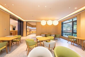 Un comedor moderno con mesas y sillas de madera, una cocina al fondo y ventanas grandes.