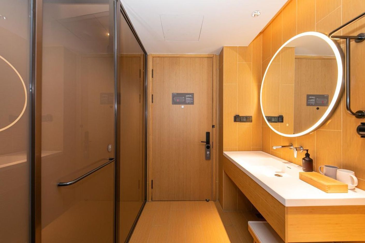 A modern hotel bathroom with a glass shower enclosure, a wooden vanity with a mirror, and a white countertop.
