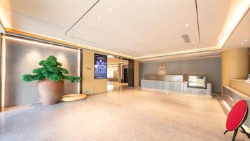 A modern hotel lobby with a reception desk, a large potted plant, and a digital information screen.