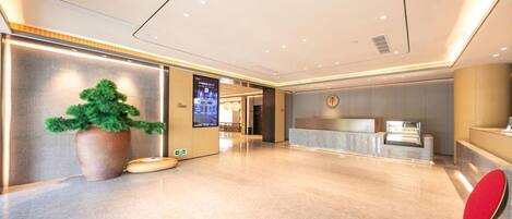 A modern hotel lobby with a reception desk, a large potted plant, and a digital information screen.