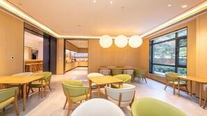 Un comedor moderno con mesas y sillas de madera, una cocina al fondo y ventanas grandes.