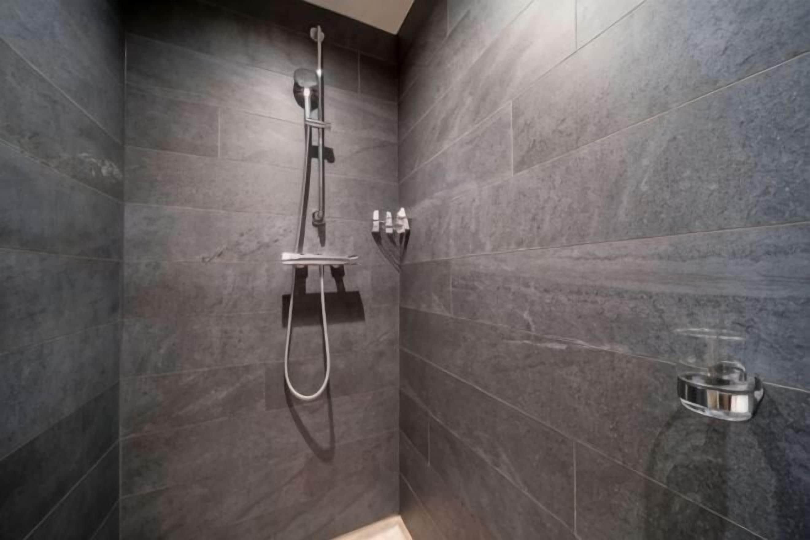 Una doccia con soffione manuale e pannello comandi a parete, in un bagno con pareti piastrellate scure.