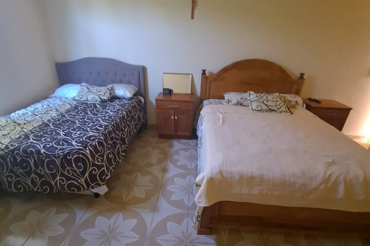 Twee enkele bedden met hoofdborden, een klein houten nachtkastje, een kruis aan de muur, en een betegelde vloer met een bloemenmotief.