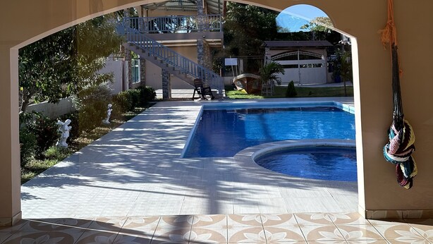 A pool area with a tiled floor, a covered walkway, and a view of a building with stairs and a flag.