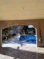 Zona de piscina con piso de baldosas, un paseo techado y vista a un edificio con escaleras y una bandera.