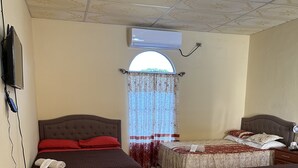 A hotel room with two beds, a TV, an air conditioner, a window with a curtain, and a trash bin.