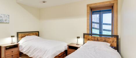 Une chambre avec deux lits simples, des têtes de lit en bois, des tables de chevet avec des lampes et une fenêtre avec des stores.