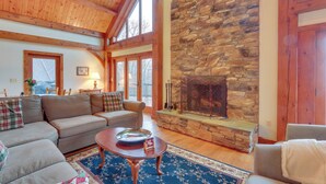 A cozy living room with a stone fireplace, wooden beams, and a large window. - Spacious Virginia Vacation Rental w/ Yard & Sauna (Purcellville)