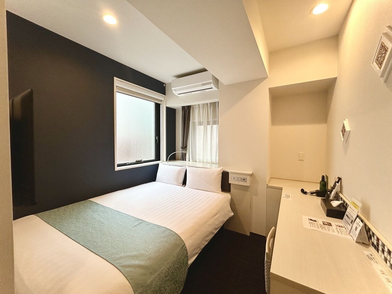 A hotel room with a bed, a desk, a window with curtains, an air conditioning unit, and a small kitchenette.