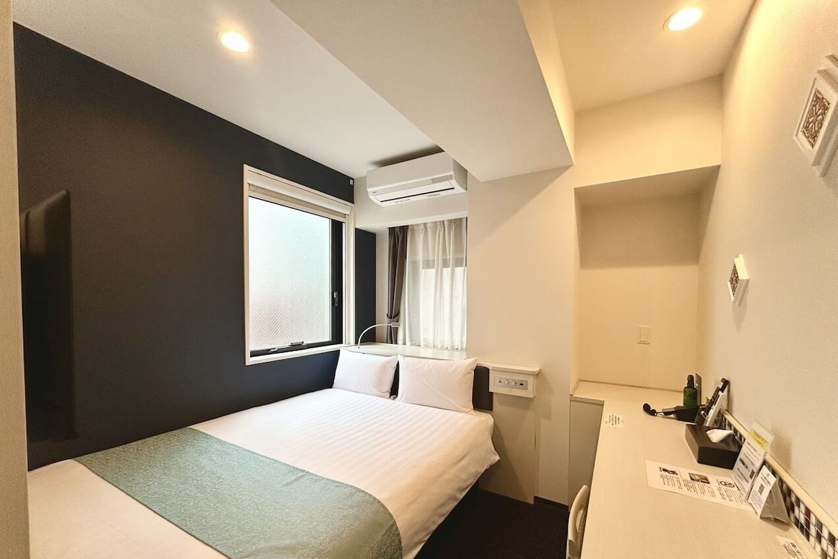 A hotel room with a bed, a desk, a window with curtains, an air conditioning unit, and a small kitchenette.