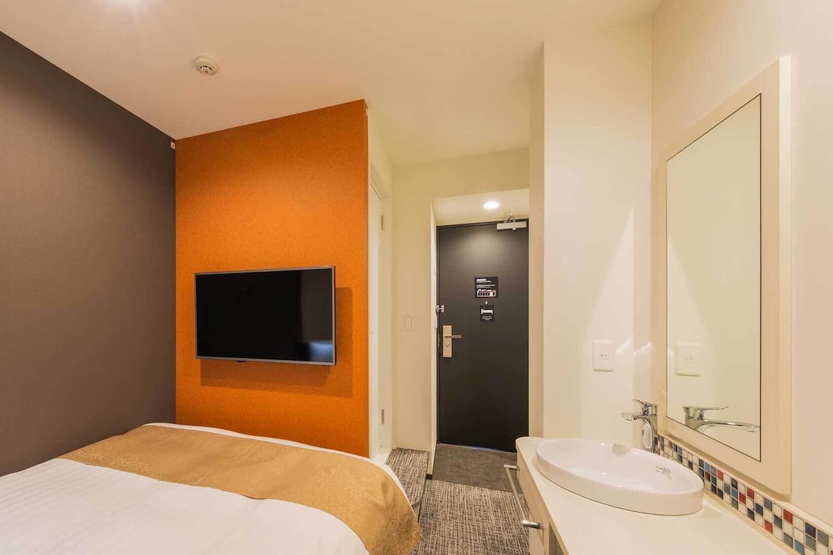 A hotel room with a bed, a mounted TV, a bathroom with a sink and mirror, and a door leading to another room.