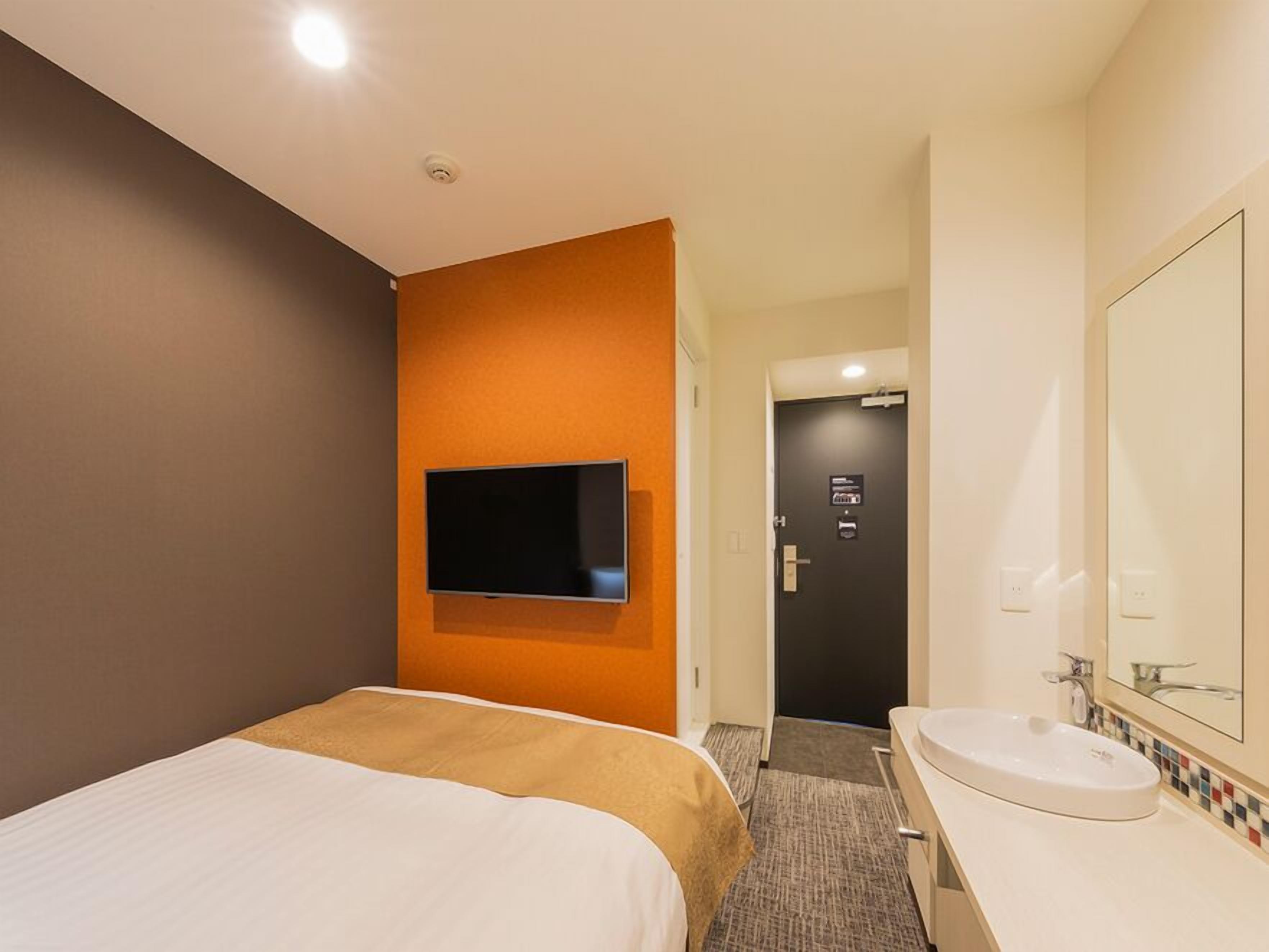 A hotel room with a bed, a television mounted on the wall, a bathroom with a sink and mirror, and a door leading to another room.
