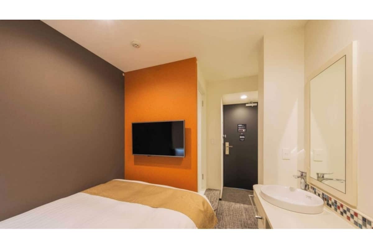 A hotel room with a bed, a television mounted on the wall, a bathroom with a sink and mirror, and a door leading to another room.