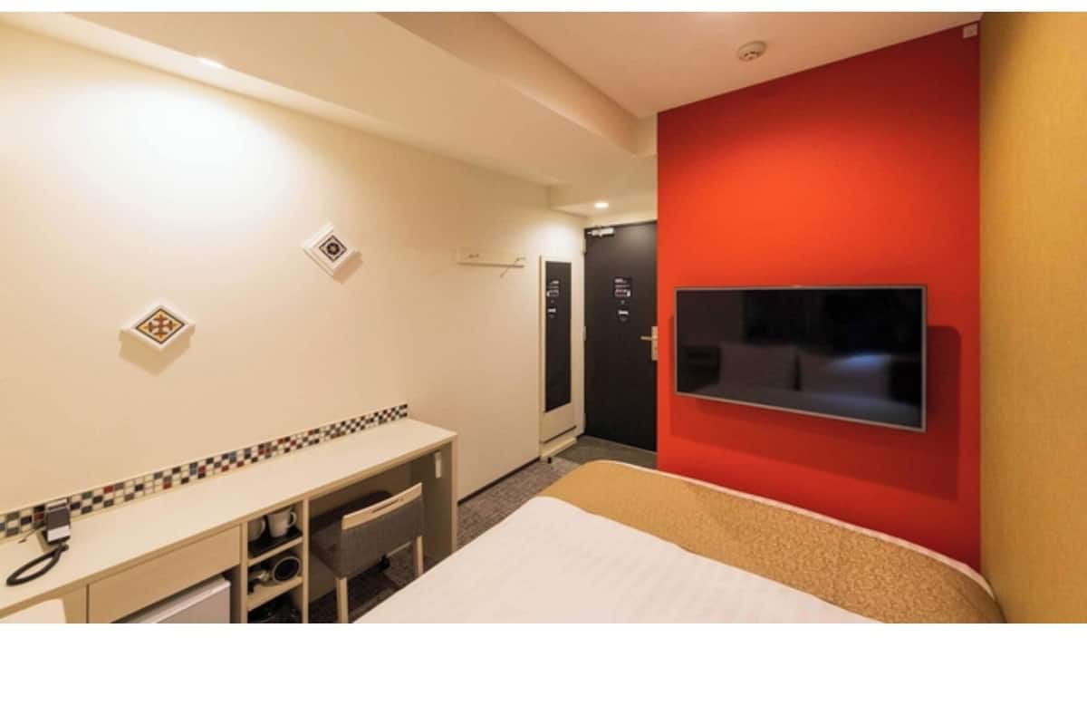 A hotel room with a bed, a desk with a chair, a television mounted on the wall, and a red accent wall.