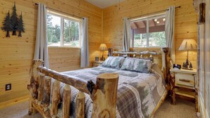 Ein Schlafzimmer in einer Blockhütte mit einem Bett, zwei Fenstern, einem Deckenventilator und einem Nachttisch mit Lampe.