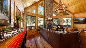 A log cabin interior with a stone fireplace, wooden walls, and large windows. - Great Escape Retreat! Hot Tub & Fire Pit! (Kane County)