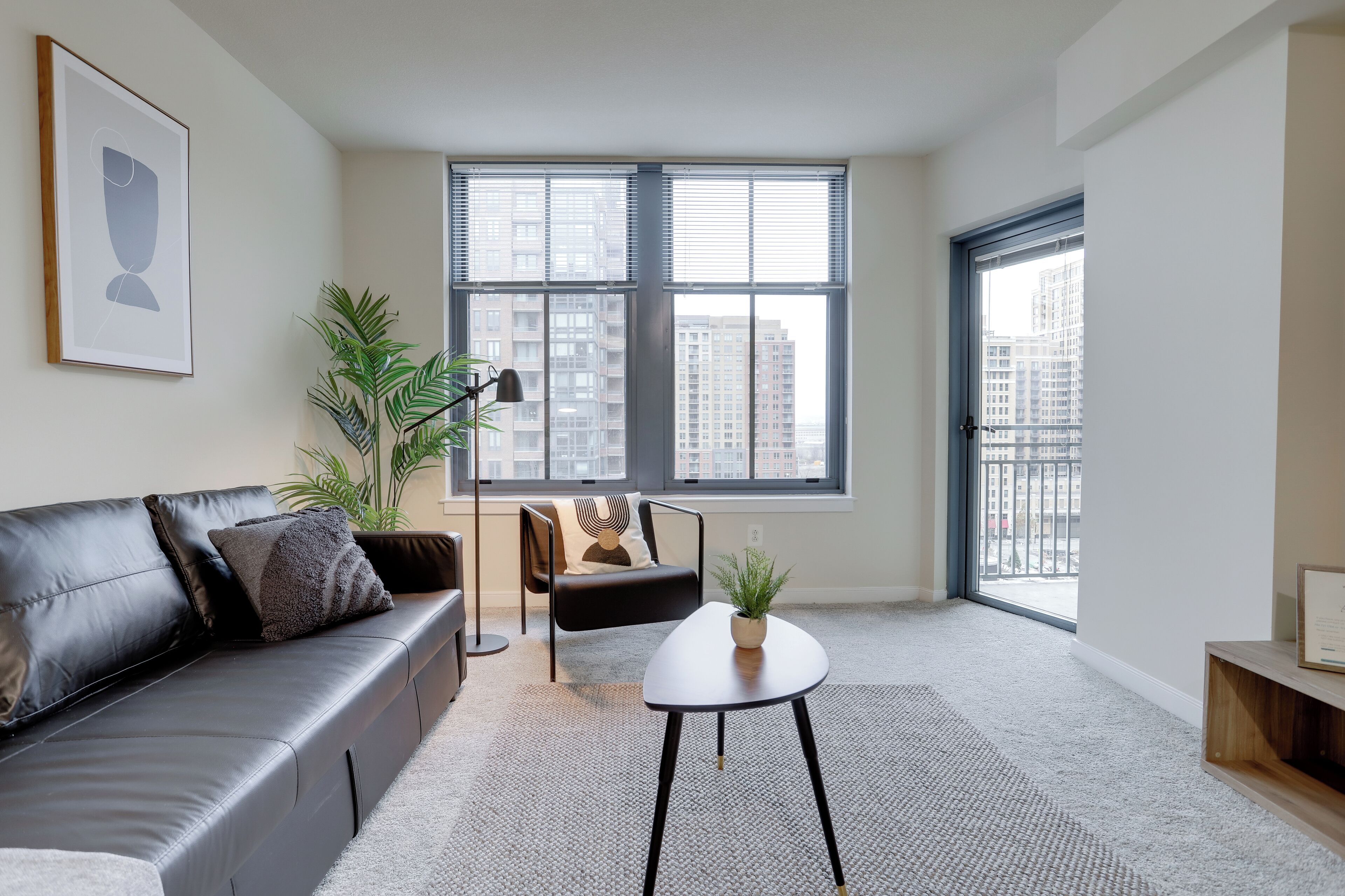 A modern living room with a black leather sofa, a small white table, a floor lamp, and a large window with blinds.