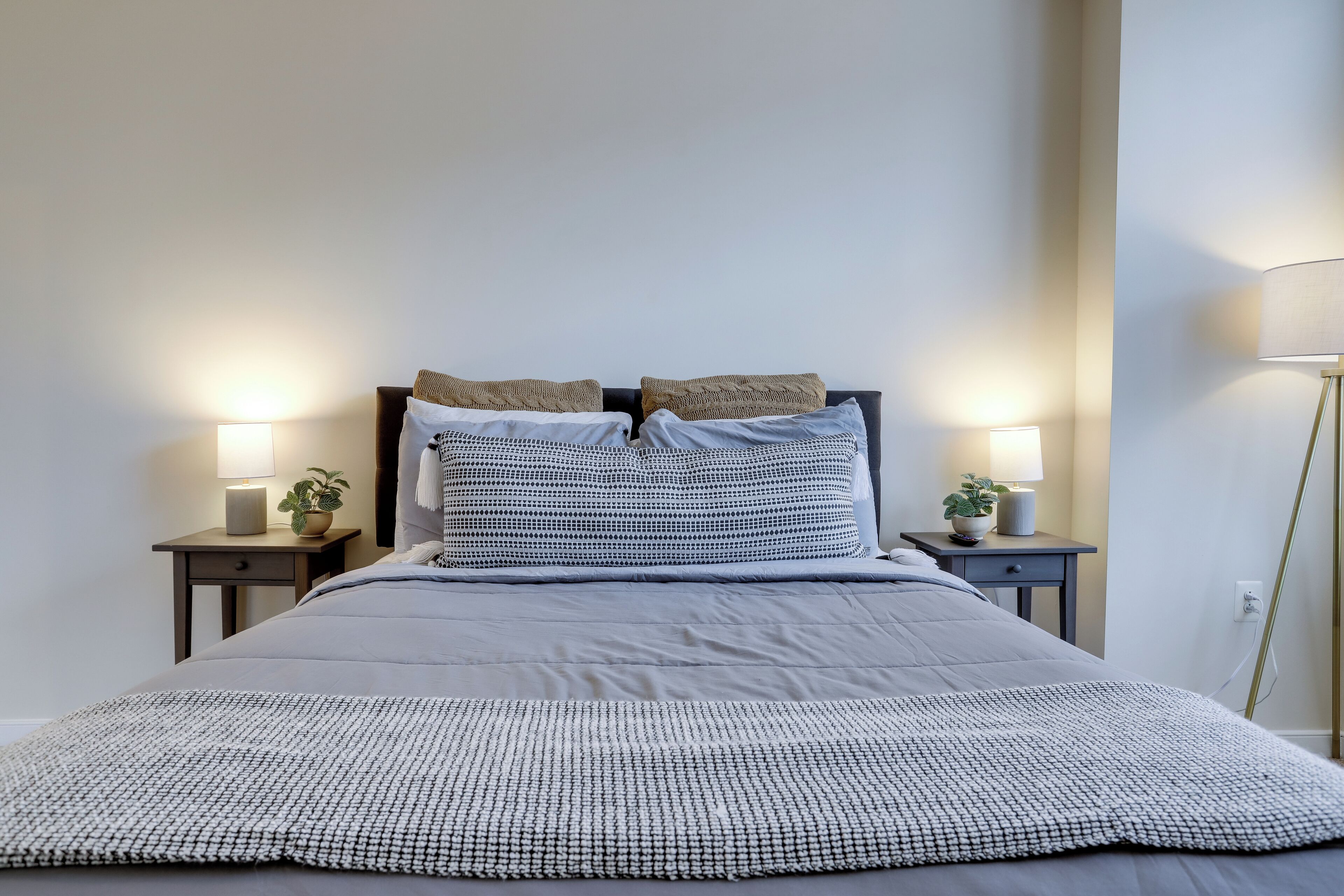 A neatly made bed with a patterned blanket, two bedside tables with lamps and small plants, and a tall floor lamp.