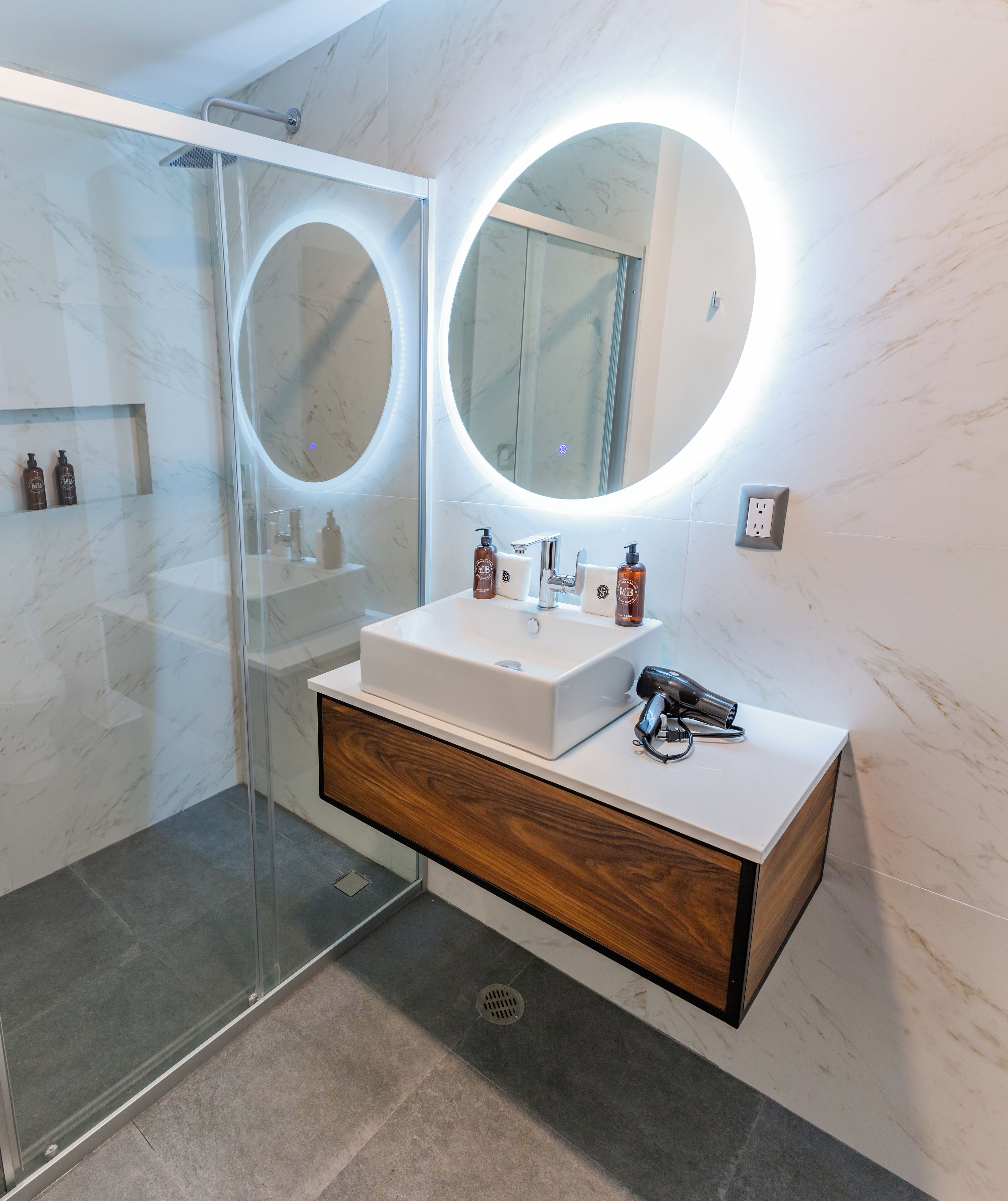 A modern bathroom with a glass shower enclosure, a white vanity with a wooden drawer, and a round mirror with a light.