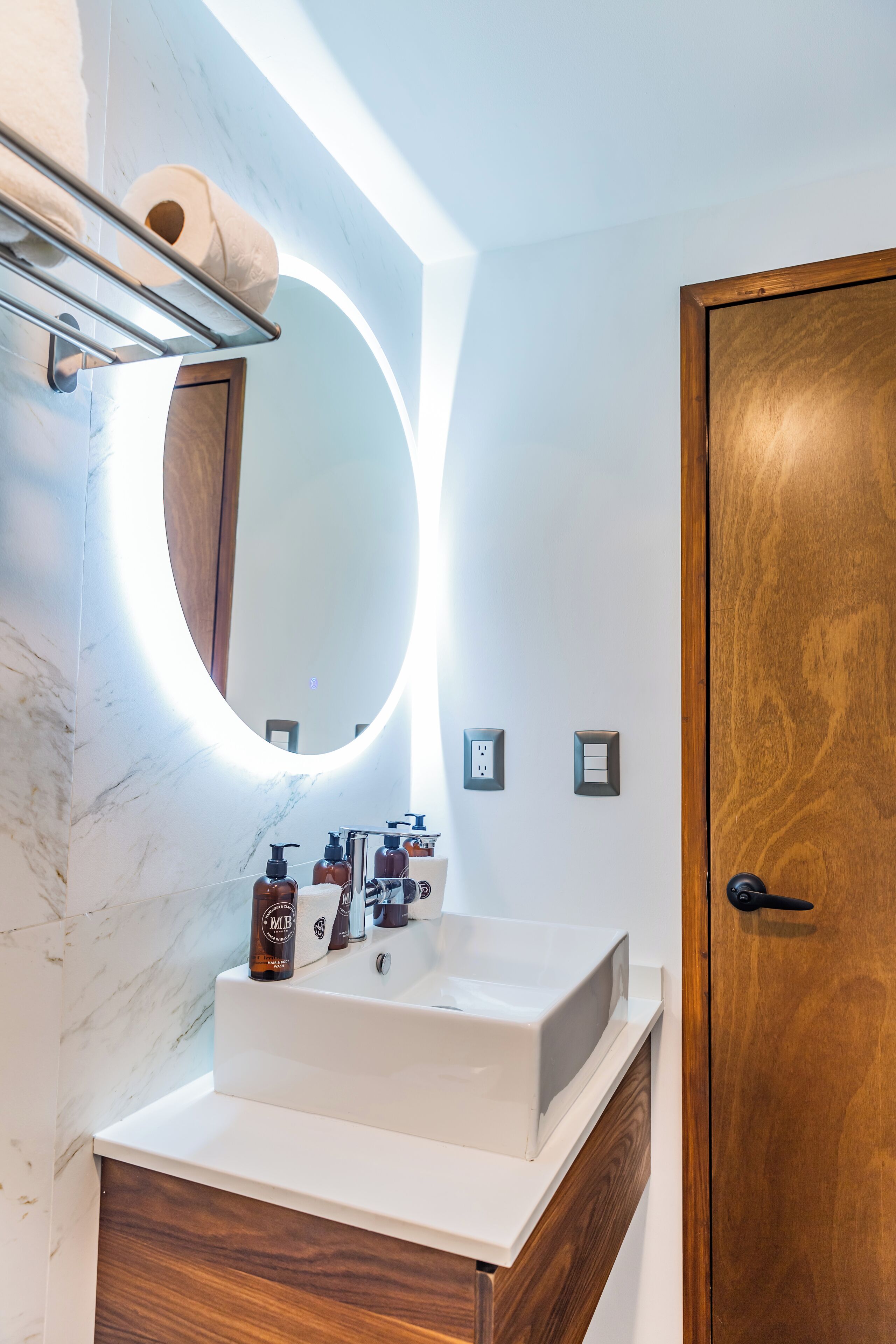 A bathroom with a white sink, wooden vanity, a round mirror with a light, and a wooden door.