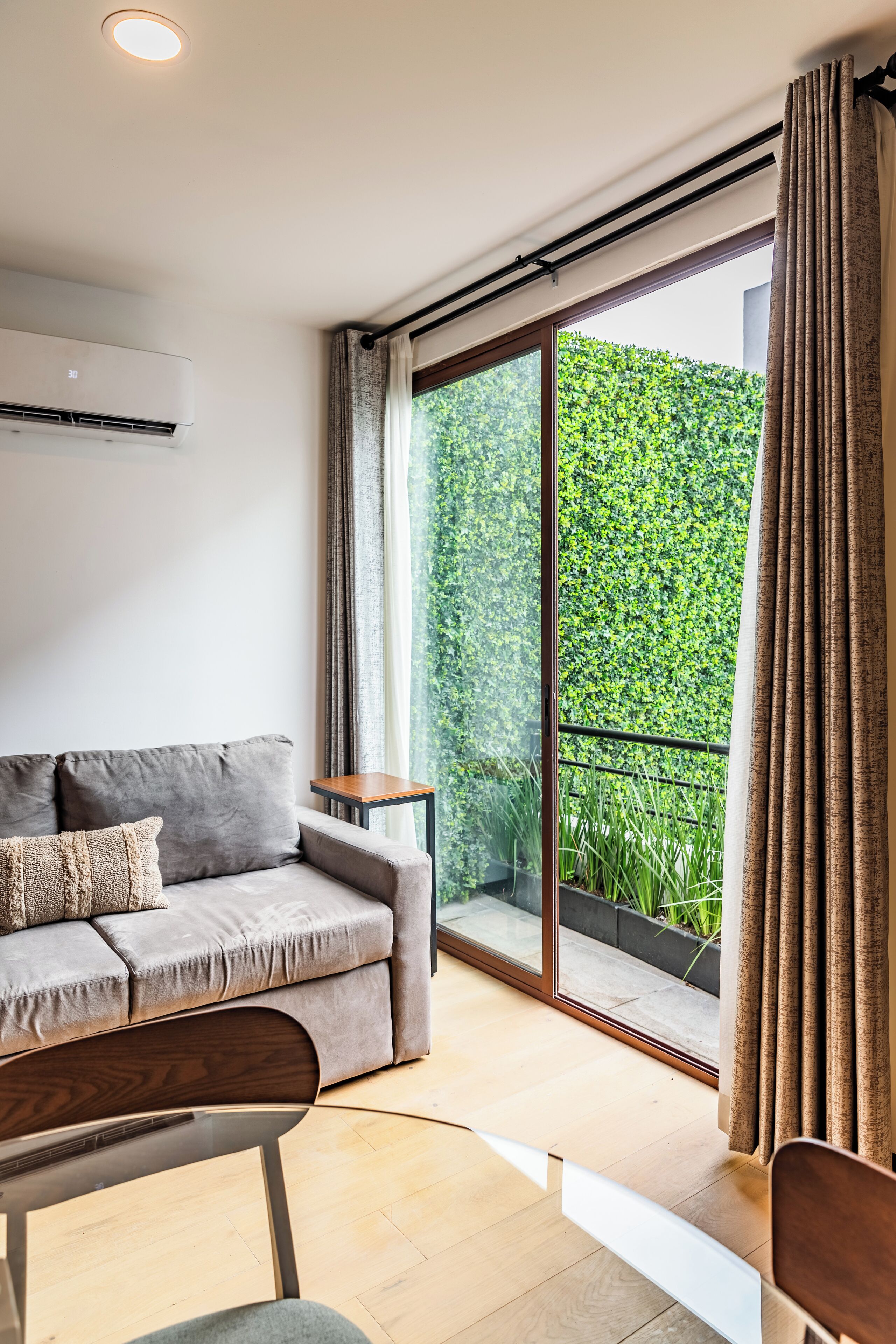 Ein modernes Wohnzimmer mit einem grauen Sofa, einem Holztisch und einer großen Glasschiebetür, die auf einen Balkon mit einer grünen Hecke führt.