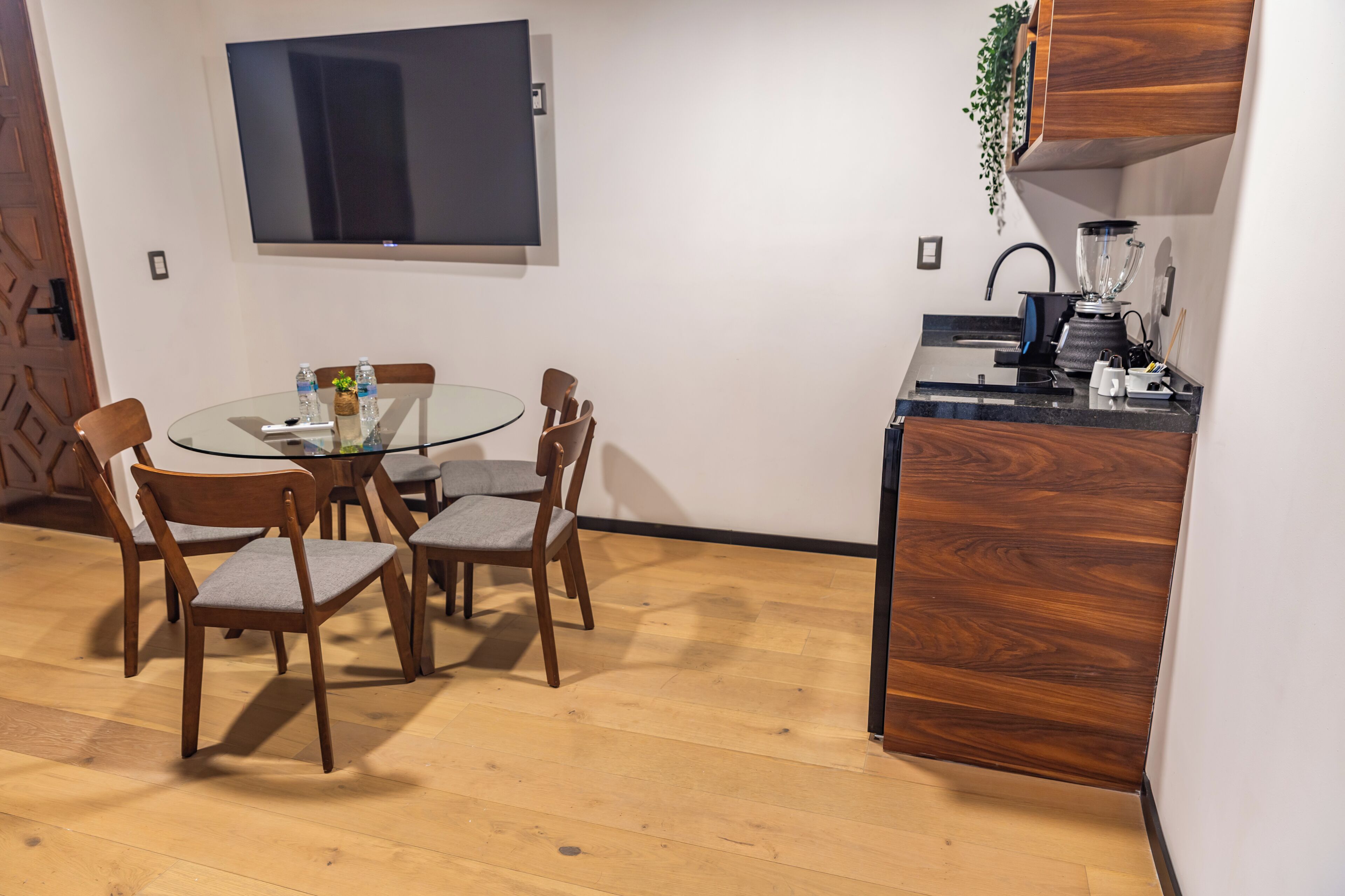 A dining area with a glass table, wooden chairs, a TV mounted on the wall, and a kitchen counter with a blender.