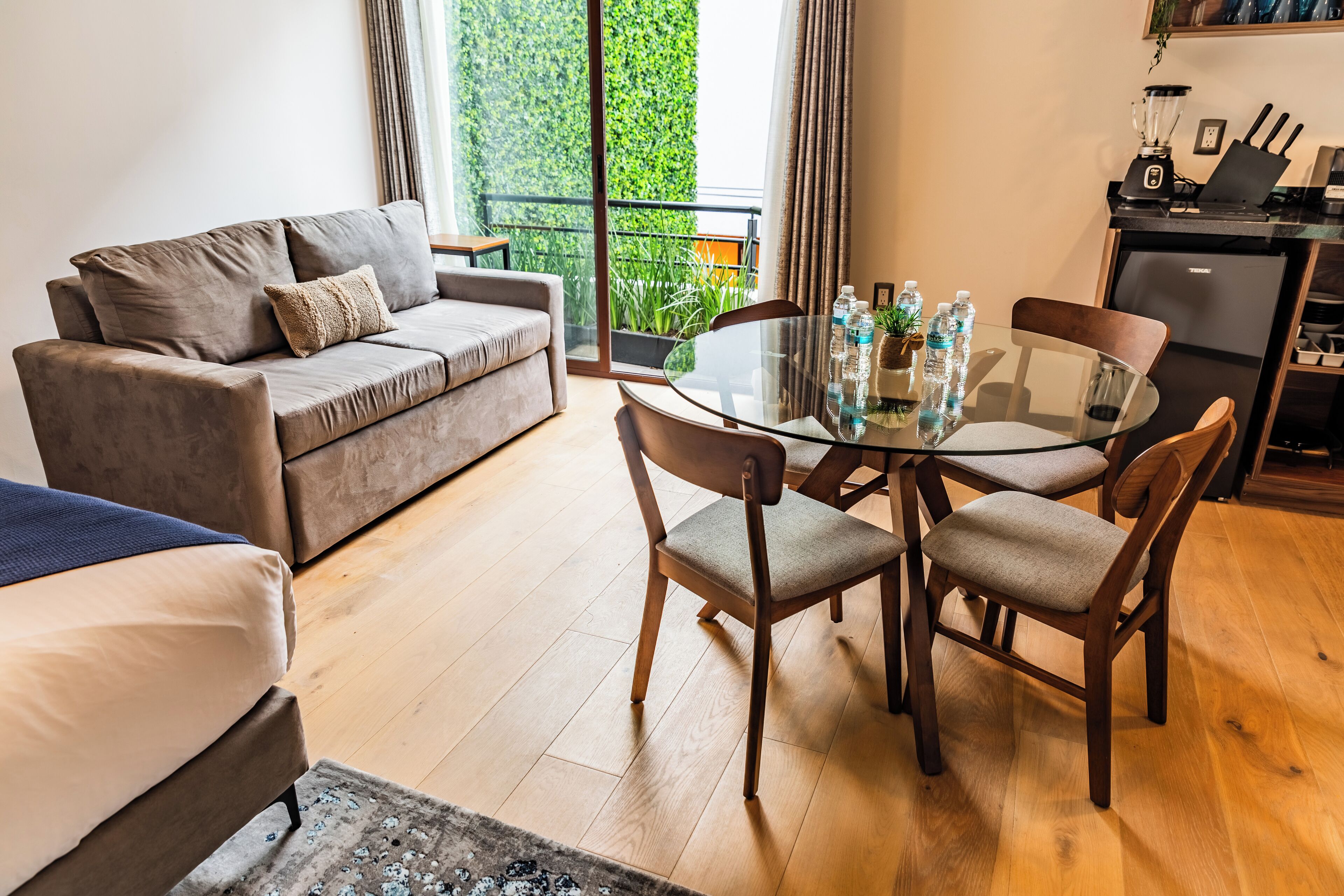 A modern living room with a grey sofa, a glass dining table with four chairs, and a kitchen area with a refrigerator and a blender.