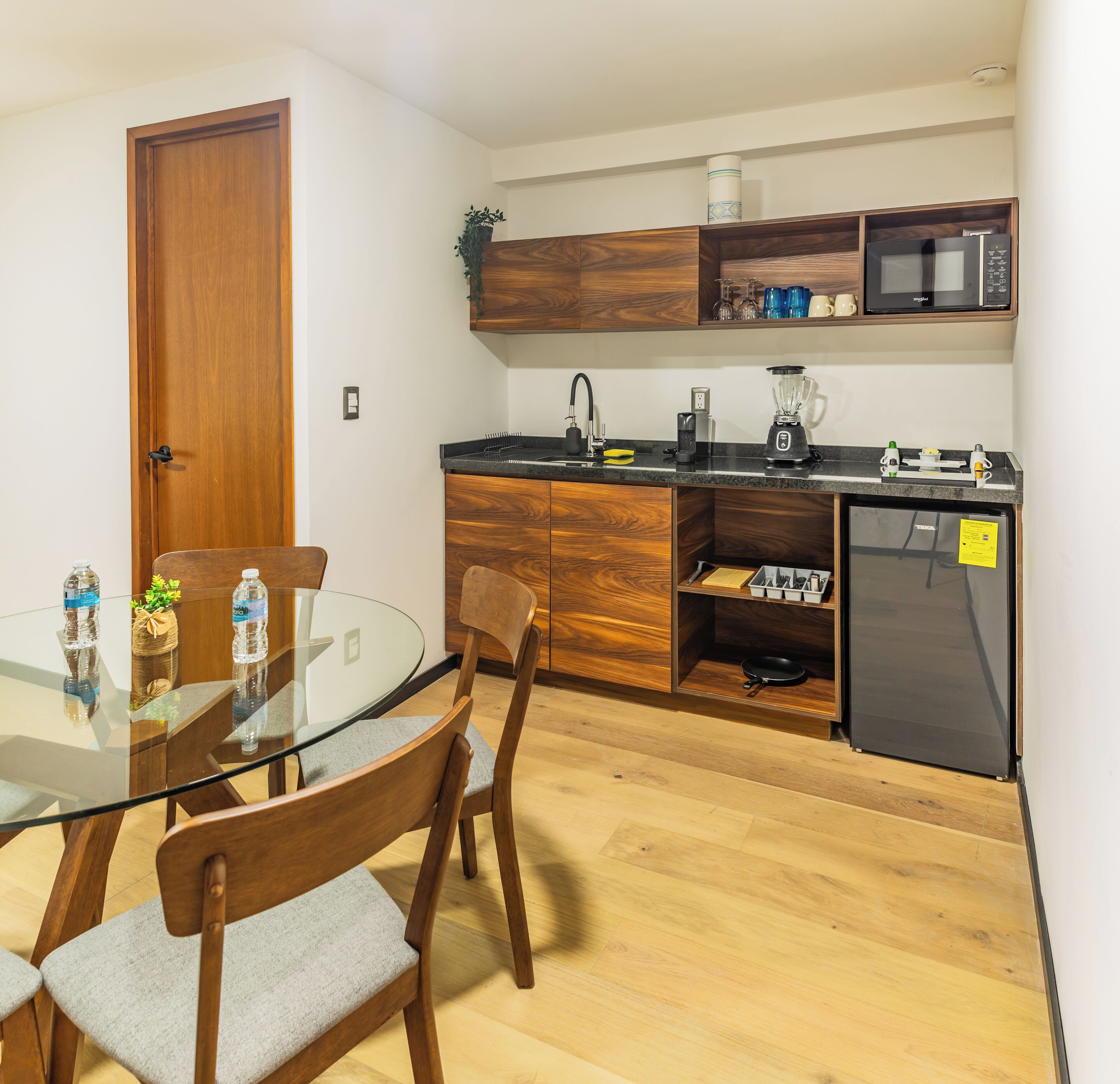 A compact kitchen with a microwave, blender, and a refrigerator. There is a wooden cabinet above the counter and a glass table with chairs.