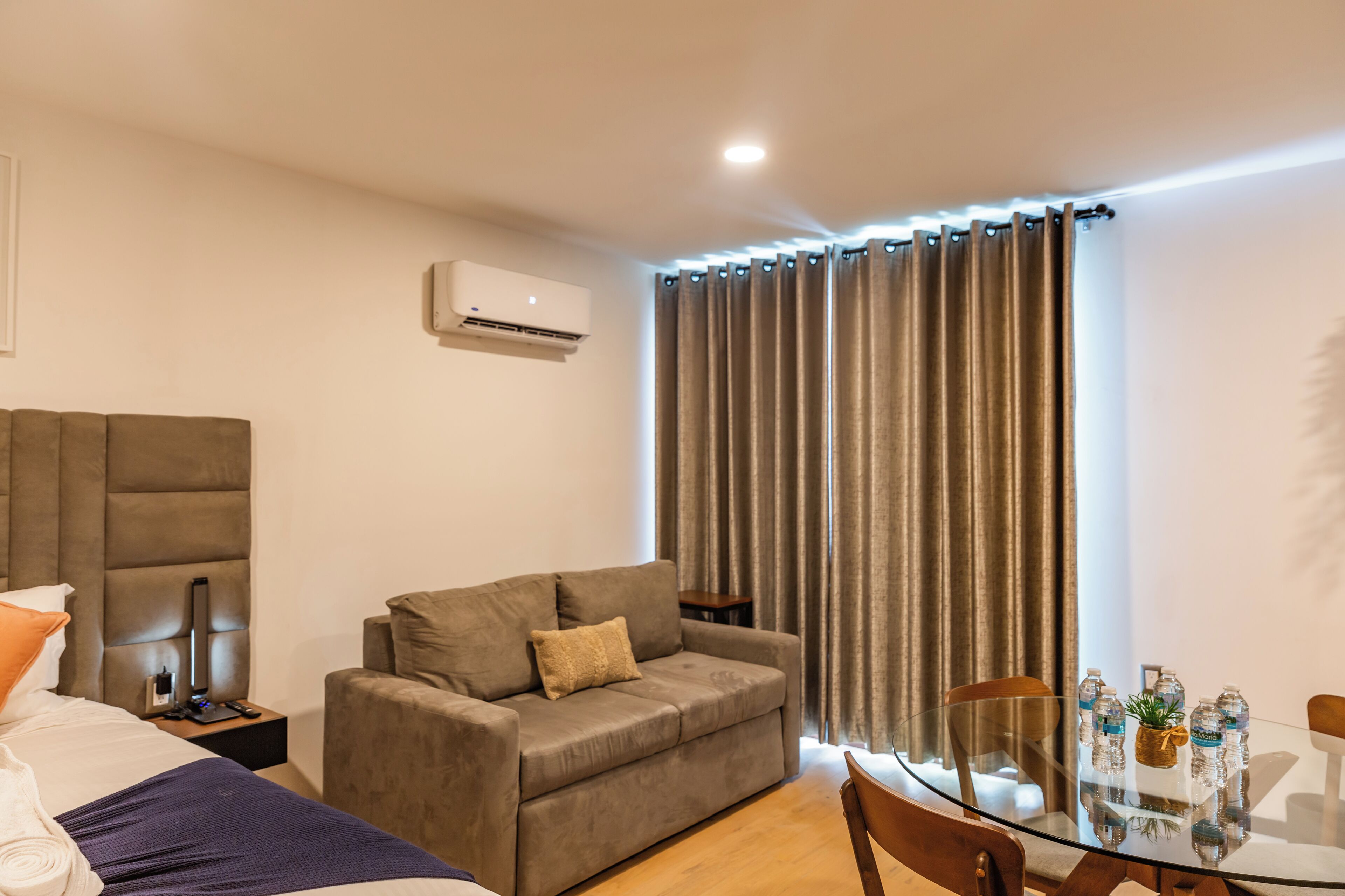 A modern hotel room with a sofa, a glass dining table, and an air conditioning unit.