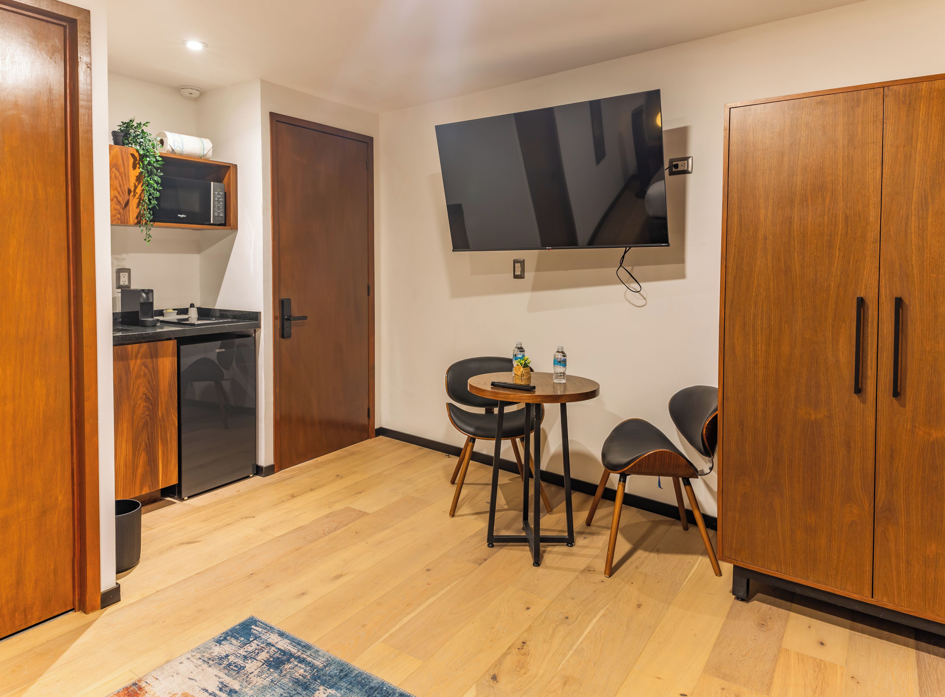 A compact living space with a kitchenette, a dining area with two chairs, a large flat-screen TV mounted on the wall, and wooden cabinets.