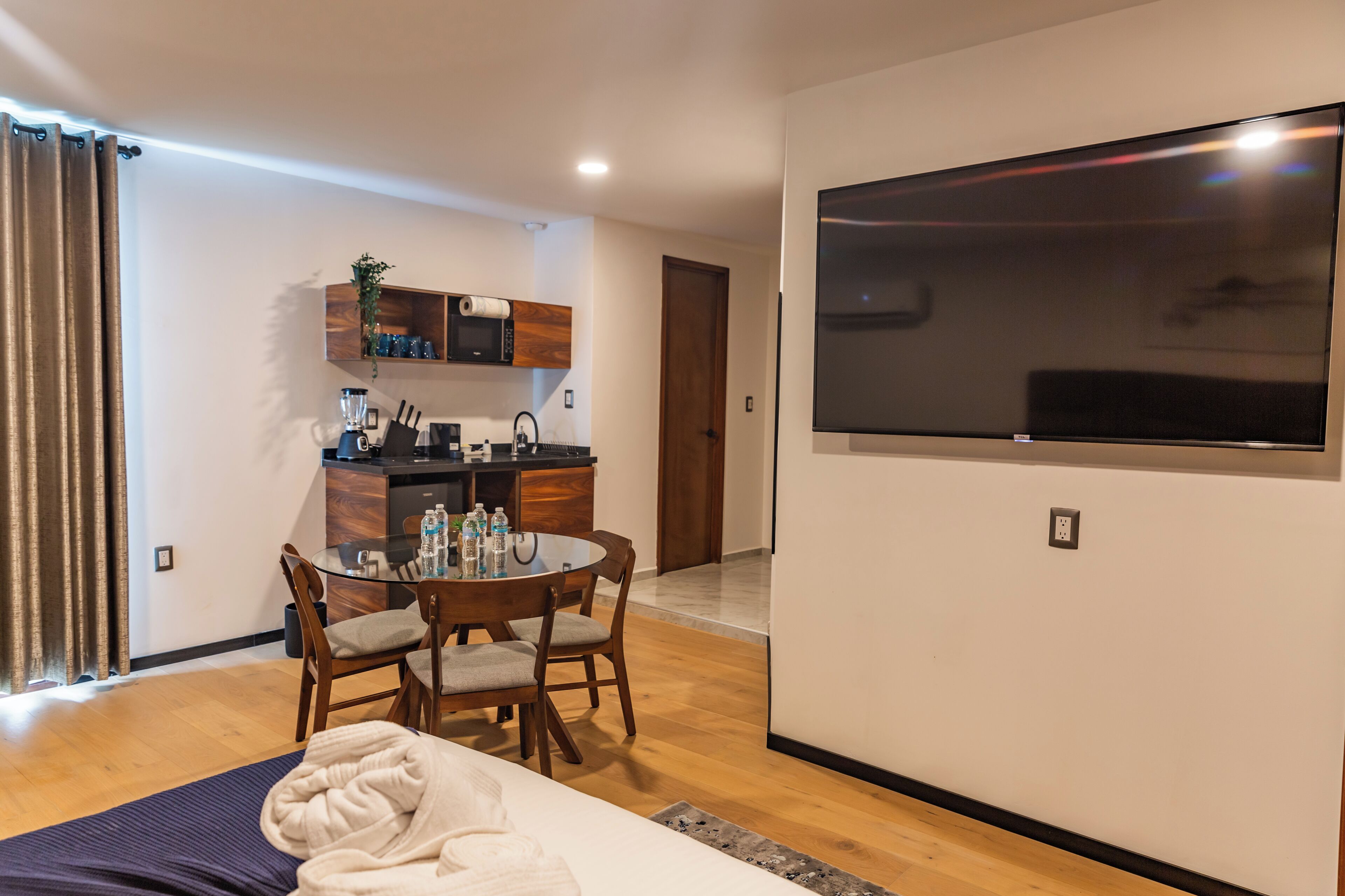 A modern hotel room with a bed, a dining table with chairs, a kitchen area, and a large TV mounted on the wall.