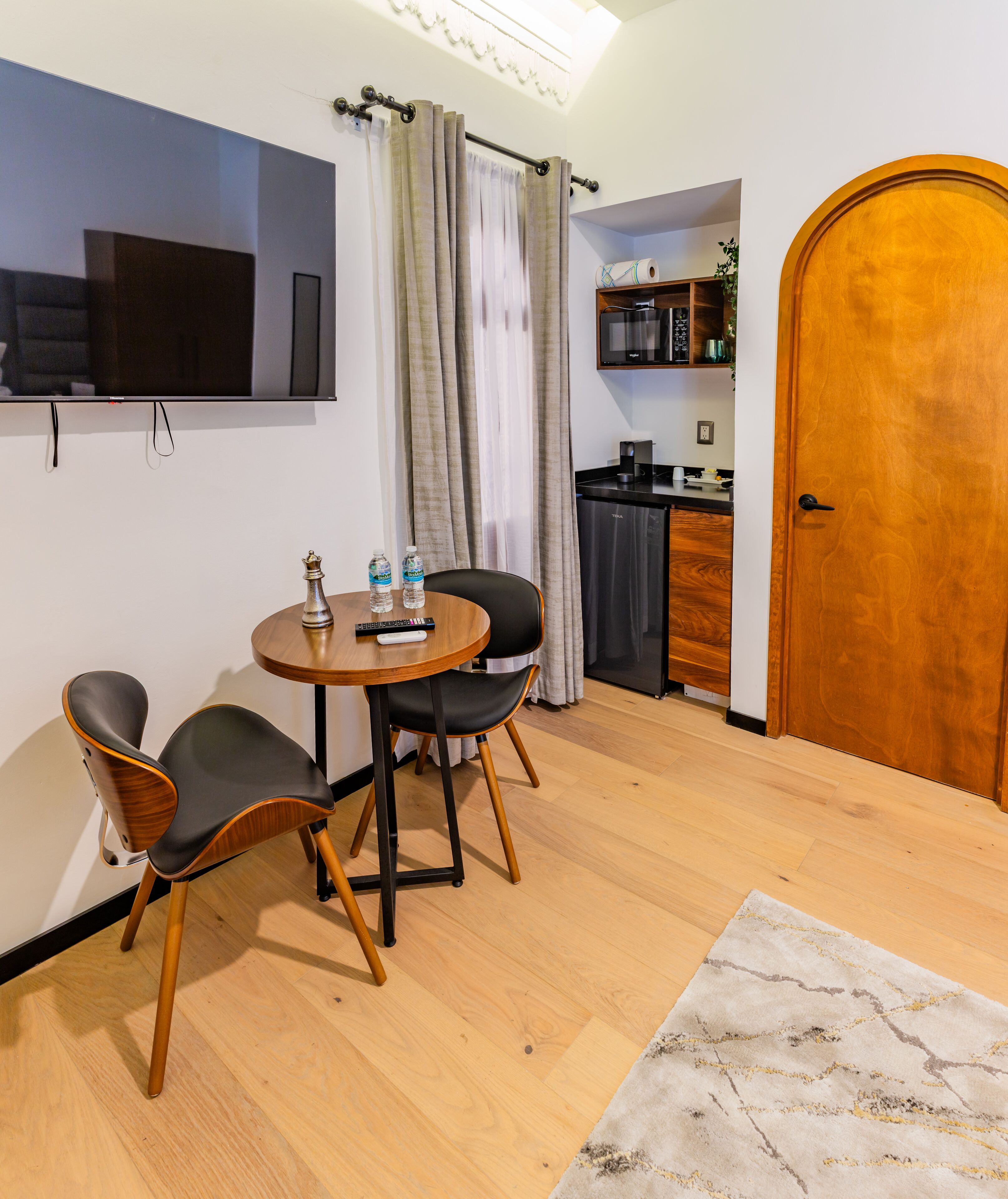 A modern living room with a round wooden table, two black chairs, a flat-screen TV mounted on the wall, a kitchen area with a refrigerator and cabinets, and a wooden door.