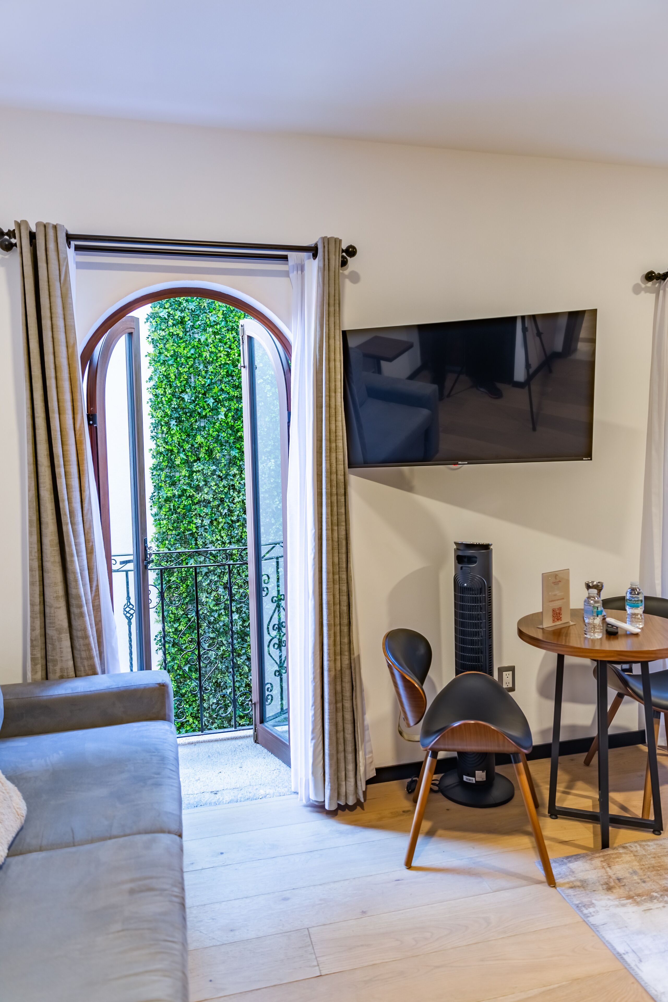 Ein Wohnzimmer mit einer Couch, einem an der Wand montierten Fernseher, einem kleinen Tisch mit einem Stuhl und einem Balkon mit Blick auf Grünflächen.